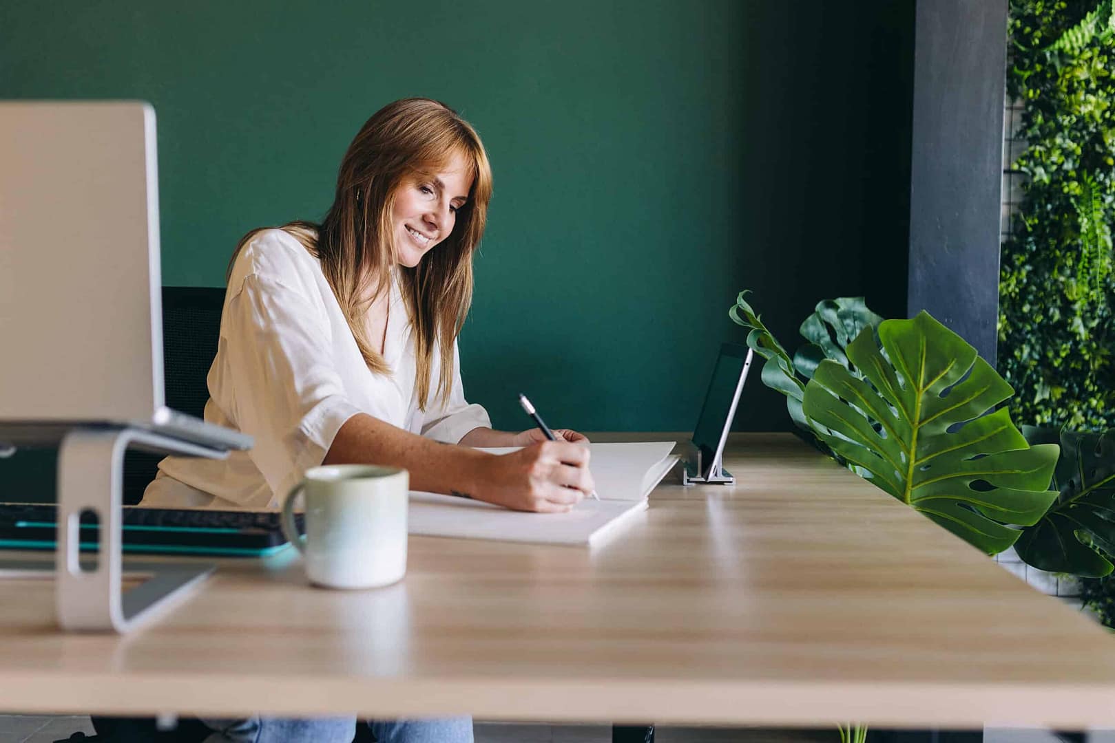 A woman smiling while writing in a notebook at a modern desk with a computer monitor and green plant decor. A smiling woman sitting at a desk writing on paper with a pen, with a laptop, smartphone on a stand, a cup, and indoor plants nearby. The wall behind her has a green section and is partially covered with vertical garden foliage.