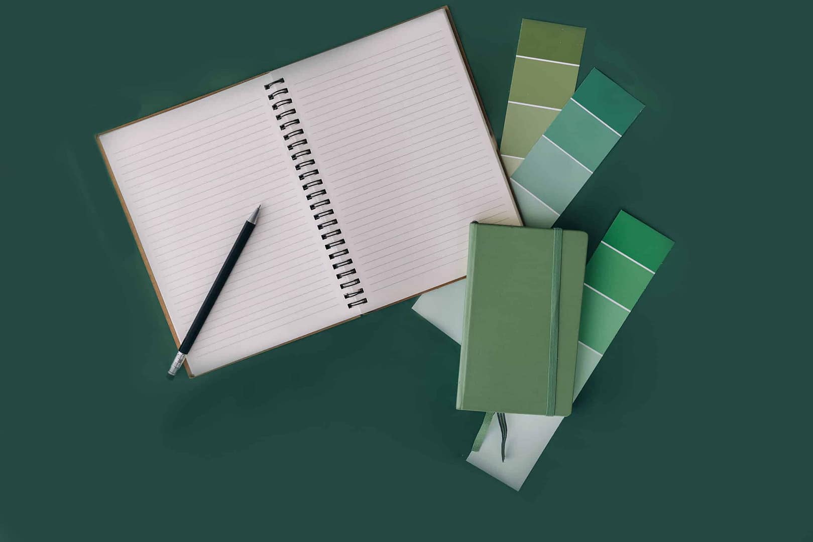 An open notebook with a pencil on it, flanked by color swatches and a closed green notebook, all arranged on a dark green surface. An open spiral notebook with a pencil on top, next to a closed green notebook and color swatches in various green shades, all arranged on a dark green background.