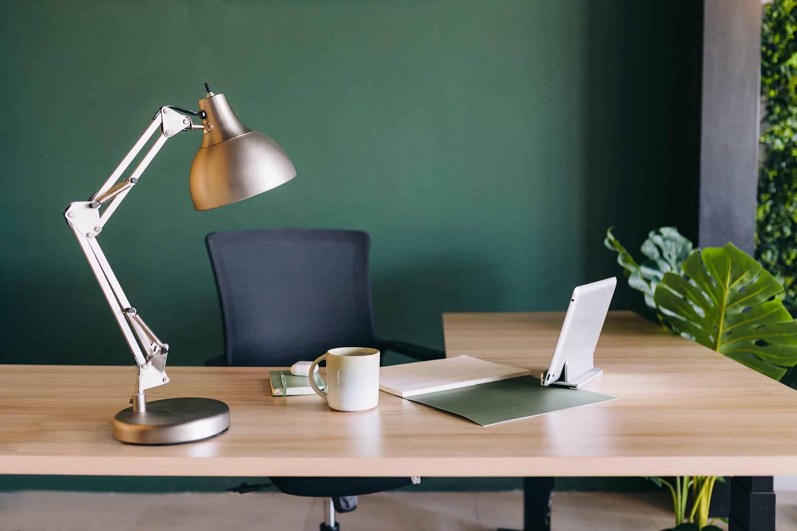 A tidy workspace with a desk lamp, laptop, notepad, mug, and a chair against a green wall. A modern workspace with a wooden desk featuring an adjustable desk lamp, a black office chair, a coffee mug, some papers, a clipboard, a tablet stand with a tablet, and a decorative plant next to a green wall with a vertical garden section.