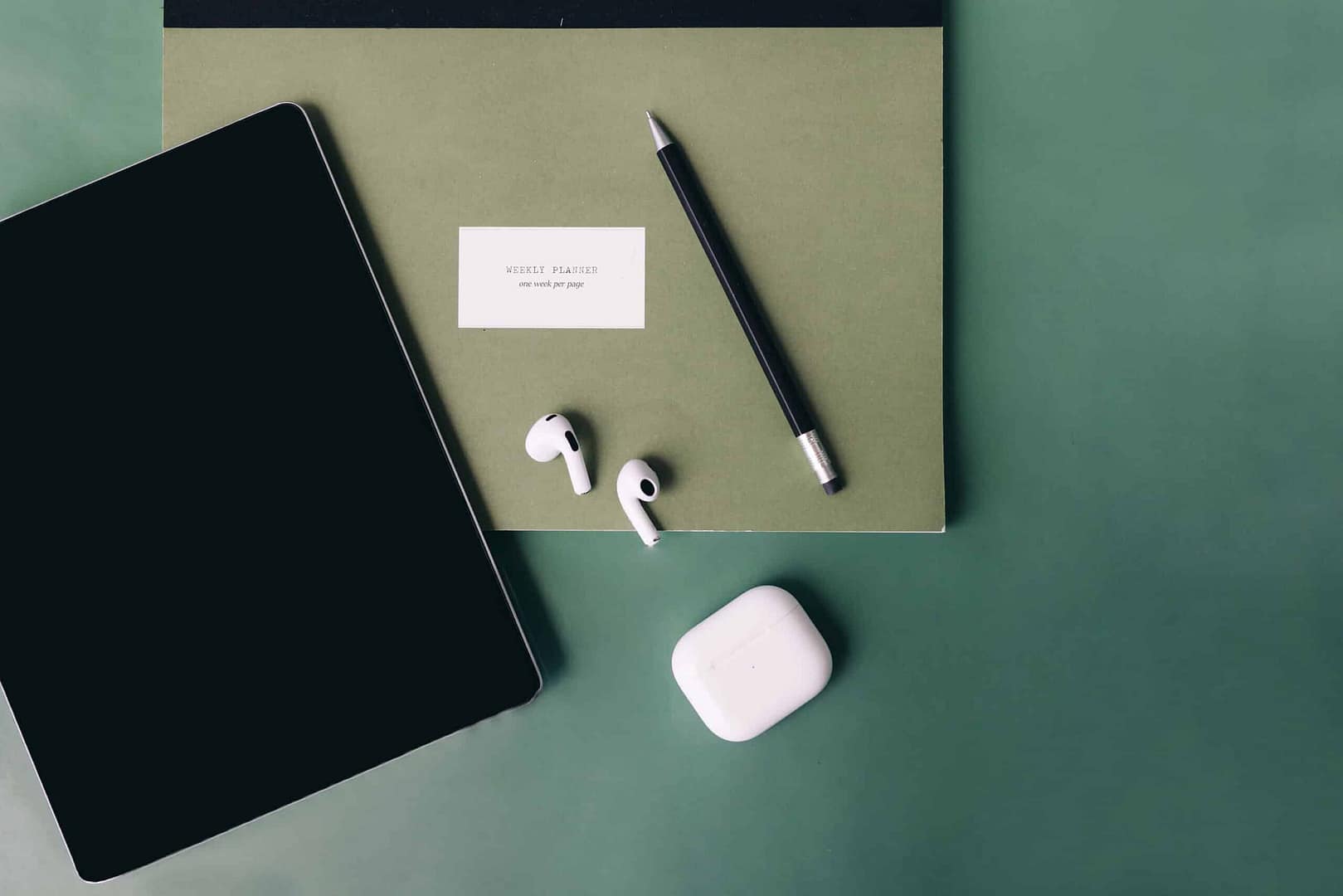Minimalist workspace with a tablet, wireless earbuds, a pencil, and a note card on a two-tone background. A tablet, wireless earbuds with charging case, a black pencil, and a card labeled 'WEEKLY PLANNER one week per page' arranged neatly on a dual-tone green and gray background.