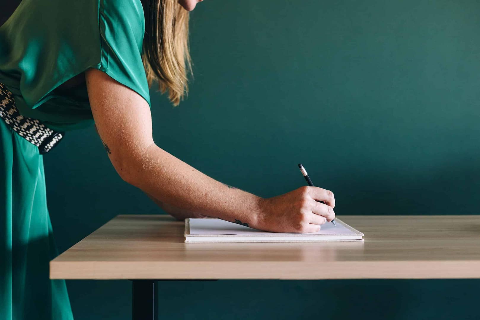 A person in a green dress writing in a notebook on a desk against a green background. A person in a teal dress is writing in a notebook on a wooden desk against a green background. Only the person's right arm and part of their upper body are visible in the frame.