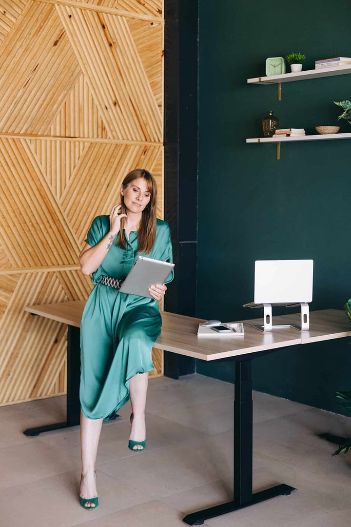 A woman in a green dress stands by a desk, talking on the phone and holding a tablet. A woman in a green dress and matching heels is sitting casually on a desk, holding a tablet and talking on the phone in a modern office with wooden geometric wall decor and minimalistic shelves.