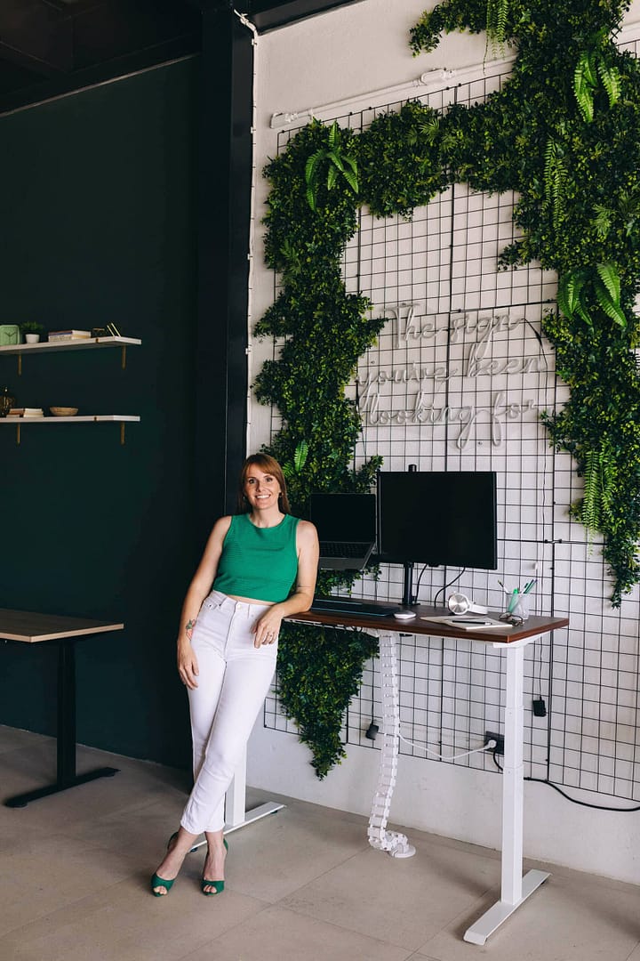 A smiling person leaning against a standing desk in a modern office with green wall plants. A smiling woman in a green sleeveless top and white pants standing by a height-adjustable desk in a modern office with green wall plants and hanging greenery.