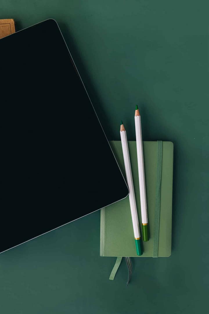 A tablet, two pencils, and a closed notebook placed on a green surface. A tablet lies on a closed green hardcover notebook, beside which are two white pencils with green erasers, on a dark green surface.