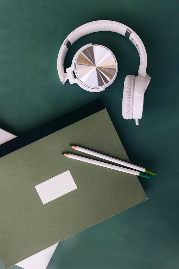 A pair of white headphones lies next to a green notebook with pencils on a dark green surface. A pair of white over-ear headphones laid on a green surface next to a dark green notebook with a blank white rectangular label attached to it and two pencils on top.
