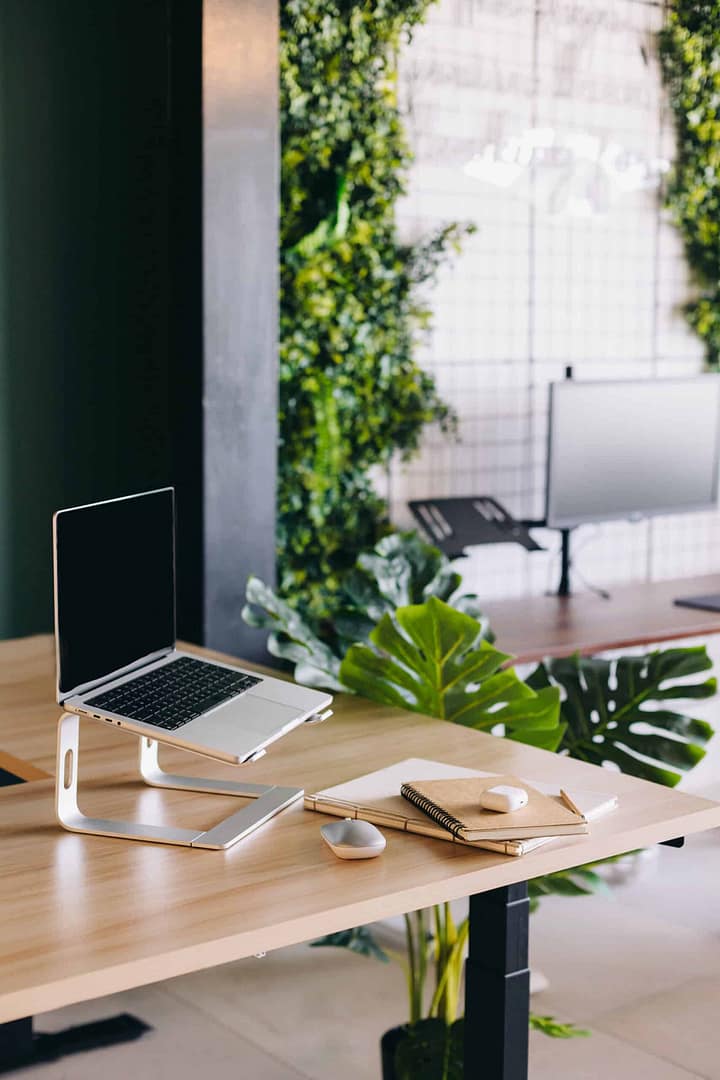 Modern workspace with a laptop on a stand, a notebook, and a mouse, surrounded by indoor plants. A modern office workspace with a laptop on a stand, a notebook, and a wireless mouse on a wooden desk. The background features indoor greenery and a blurred view of a whiteboard.