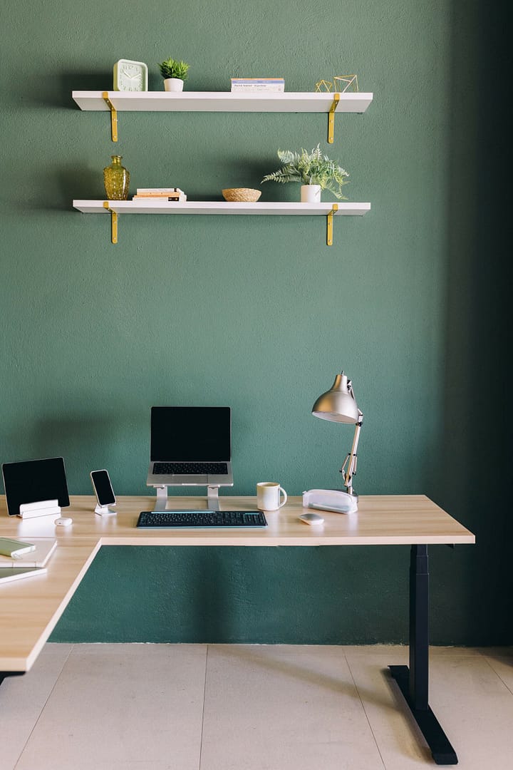 A modern home office setup with a tidy desk, laptop, desk lamp, and decorative shelves against a green wall. A minimalist home office setup with a light wooden desk against a green wall, equipped with a laptop raised on a stand, a tablet, a smartphone, a desk lamp, a cup, and some stationery. Two white shelves with decorative items and plants are mounted above the desk.