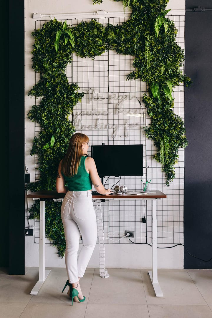 Woman standing at a desk with a computer monitor, against a wall decorated with green plants. A woman in a green sleeveless top and white pants stands at a standing desk with a computer monitor, facing a grid wall with lush green foliage. She is viewed from the side and appears to be working or studying.