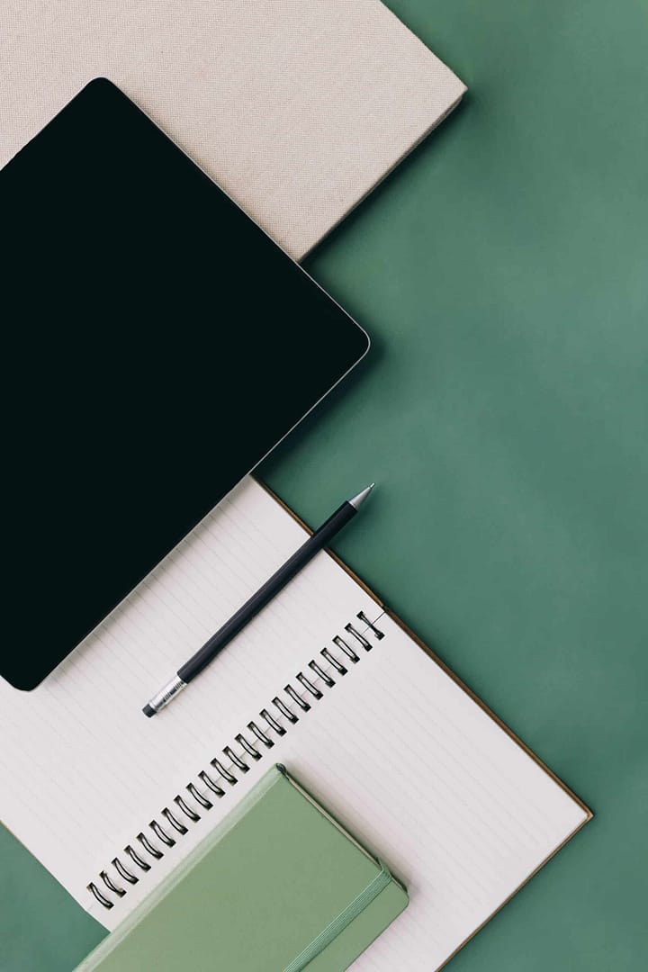 A tablet, a pencil, and notebooks arranged neatly on a green surface. A flat lay of a workspace with a tablet, a notebook with a pen on top, and a closed planner on a green background.