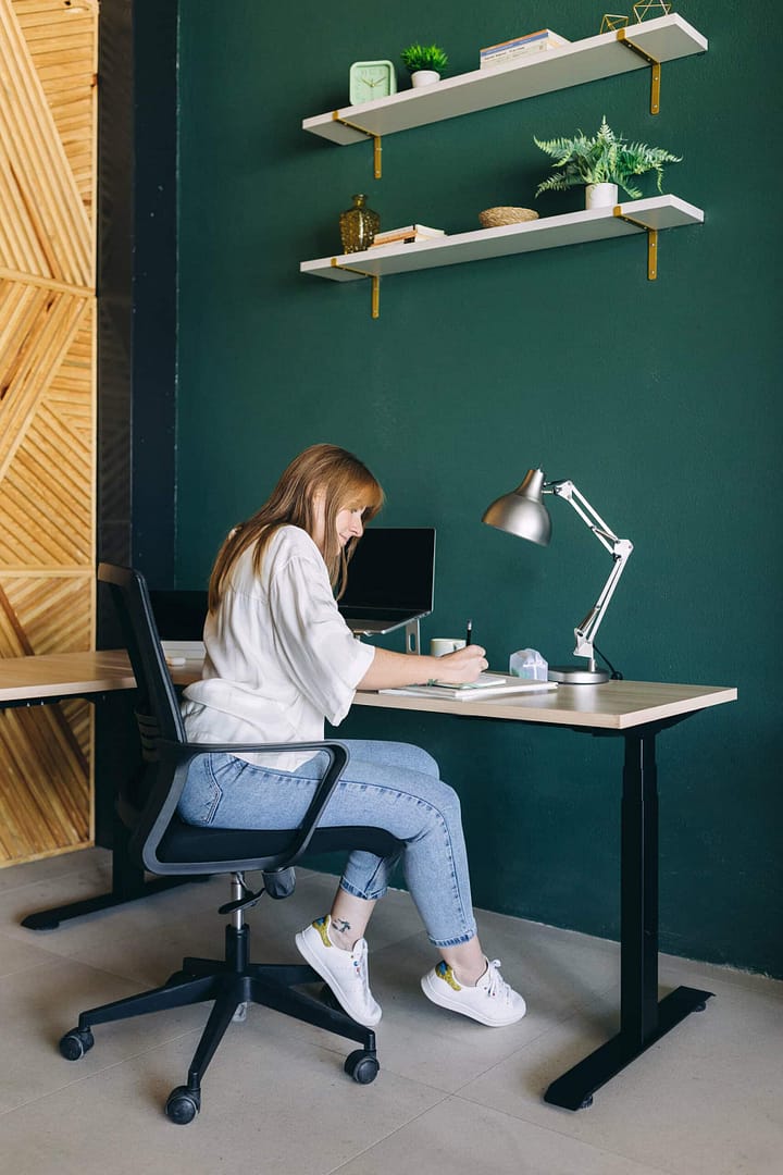 Woman working on a laptop at a modern home office desk. A woman sits at a desk in a modern home office, working on a laptop. The room features a dark green wall, floating shelves with plants and decorative items, and a wooden herringbone pattern on an adjacent wall. The woman is wearing a white shirt, blue jeans, and white sneakers, focusing on her notebook with a desk lamp beside her.