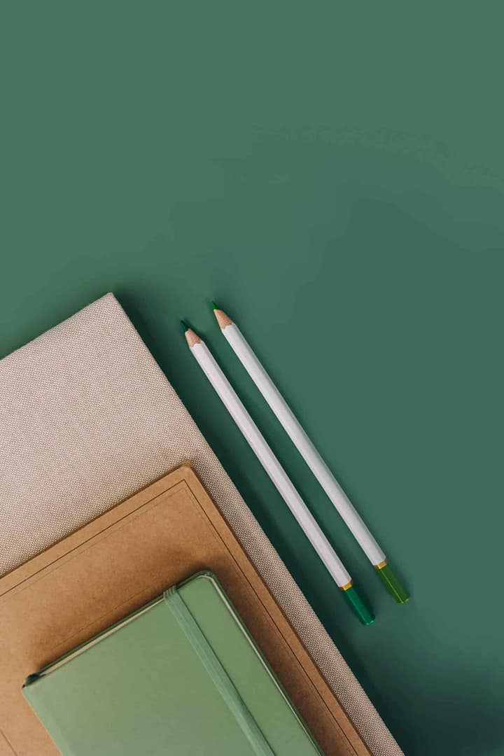 Two pencils on a green background with two closed notebooks. Two green-tipped white pencils placed on a green backdrop next to a stack of notebooks with a brown top notebook and a green one beneath.